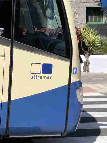 Part of a blue and yellow coach with the word "Ultramar" near a crosswalk with a background of a building and plants.