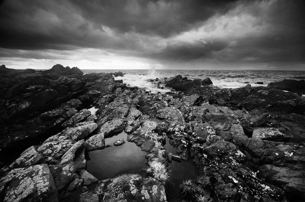 Black and white photo of a rocky foreshore leading out to the sea, with dark clouds above.