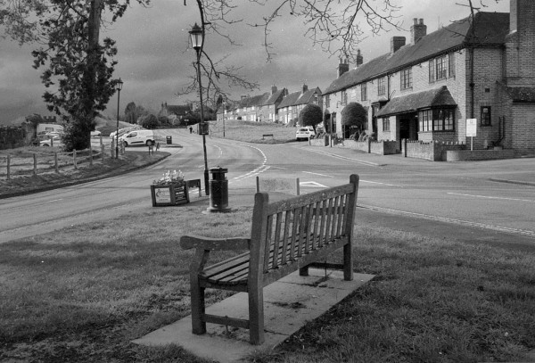 Black and white photo showing a wooden bench in the foreground, behind it an old-fashioned street lamp and a waste bin. Two roads come fgrom left and right to meet and continue up through the image with a grassy area to its right. At the right hand edge is a public house, beyond it a strip of cottages. At the left is a conifer and a car park.