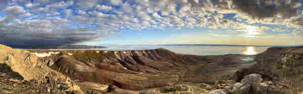 A panorama from a top of a wide sweeping valley that leads down to the Arctic Ocean. The sides of the valley are fluted with gullies and richly coloured in browns and greens. Some snow remains a long the edges of the valley. The blue sky is dotted with small clouds and the low Sun casts a golden glow along the horizon. Ice remains on the ocean. 