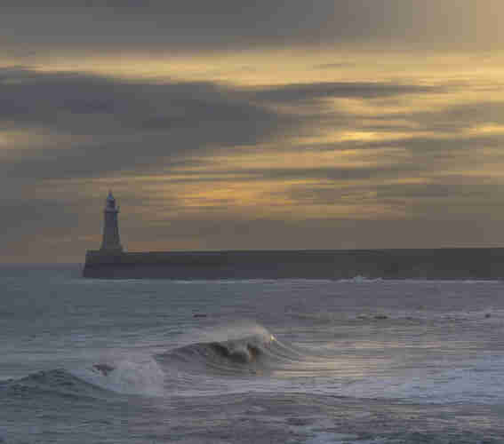 A breaking wave catches the sun. Behind a sea wall and lighthouse.