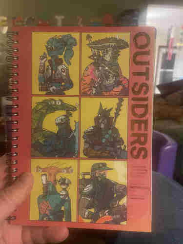 A person holding a spiral-bound notebook featuring a cover with a grid of illustrated sci-fi characters, titled "OUTSIDERS."