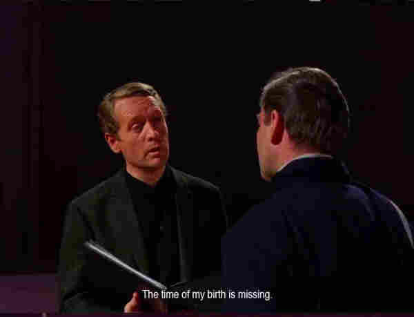 A man in a black jacket and shirt is speaking to another man. The first man is holding a folder and saying, “The time of my birth is missing.”
