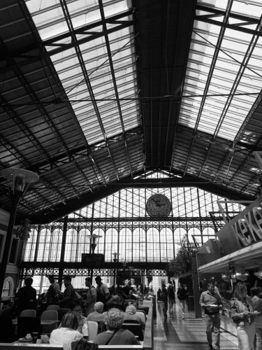 My photo with a black and white filter of the inside of the station which is like a huge warehouse with windows at the ends and in the roof. There is people and a fast food restaurant in the bottom of the photo and the windows and a huge clock behind