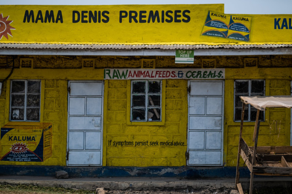 A brightly painted yellow building with signage for “MAMA DENIS PREMISES”, advertisements for various products, including “RAW ANIMAL FEEDS & CEREALS”, and the mention on the wall “If symptoms persist, seek medical advice”
