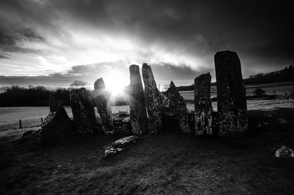 Black and white photo of the sun setting behind the tall, standing portal stones of Cairnholy Chambered Tomb.