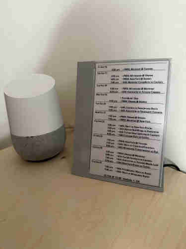 Image of an epaper display with a grey bezel on the left and bottom propped up next to a white and grey google home device. The display shows a list of upcoming sporting events.