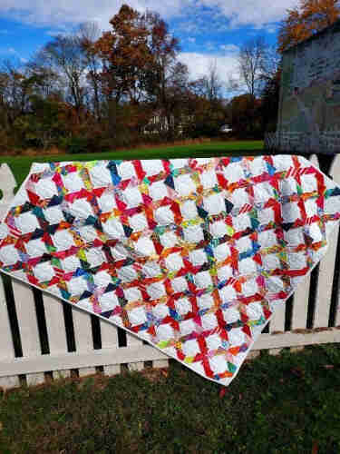 a lattice quilt, multi-colored bars on white background, criss-crossing, the quilt is on a fence, blue sky behind, autumn, the trees leafless, some with orange leaves 