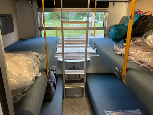 Photo of the inside of a sleeper train compartment. Two blue bunks visible either side of a ladder…two upper bunks out of frame. There is a window behind the ladder. Bags and blankets are piled up on one bunk on the right.