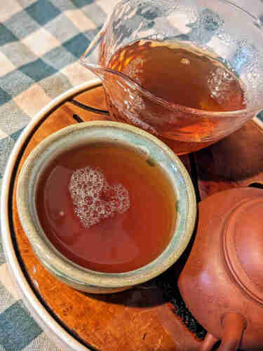 A gongfucha setup. The teacup is filled with tea and a heart made out of many small bubbles is floating on the surface. 