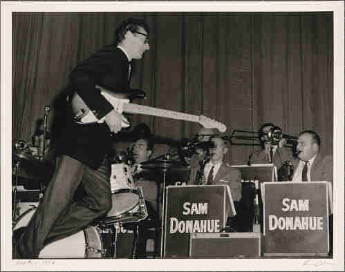 Buddy Holly runs across the stage holding his Fender guitar under one arm with Sam Donahue and his Orchestra playing behind him. January 19, 1958; Auditorium Theatre, Rochester, NY.