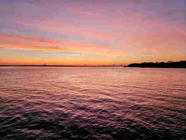 View across a vast waterway at sunrise. The blue sky is swept with bands of thin whispy clouds. The rising sun casts bands of deep yellow and orange across the horizon, making silhouettes of bridges and shipyard cranes. The colors bounce off the clouds in shades of pink and purple.