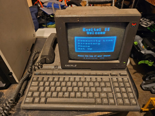 An EXELTEL II communications terminal sitting in a garage. It has a monochrome CRT, built in QWERTY keyboard and telephone handset. 