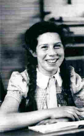 A smiling girl sitting on a school bench. She put her hands on the table, on which you can also see a notebook. Her hair is long - the braids fall to her shoulders. She is also wearing a short-sleeved blouse. A smile reveals her upper teeth.