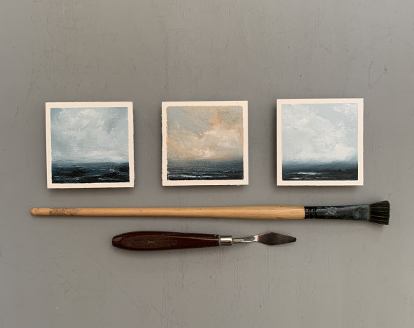 Three original seascape oil paintings by Tisha Mark, "Soothing Sea 1-3" oil on Arches oil paper, 3"x3' (2024). Three tiny seascapes arranged in a row with a paint brush and a palette knife placed below them for scale. The two on the ends are painted in a limited palette of blues and whites. The one in the middle has grayish-blue and orange cloud formations over a dark blue ocean.