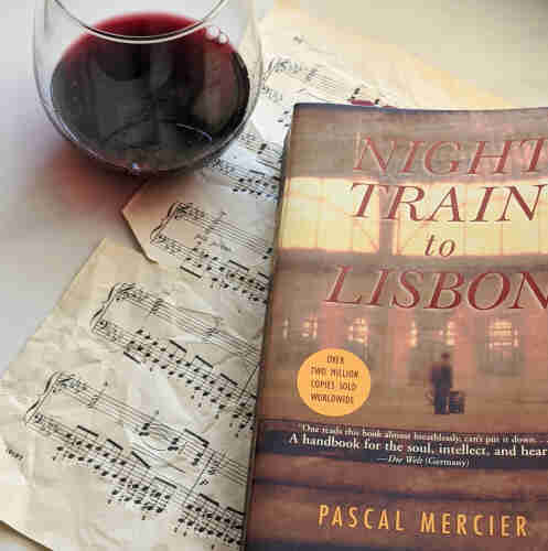 A small glass of red wine, I piece of sheet music, and the book, Night Train to Lisbon by Pascal Mercier.