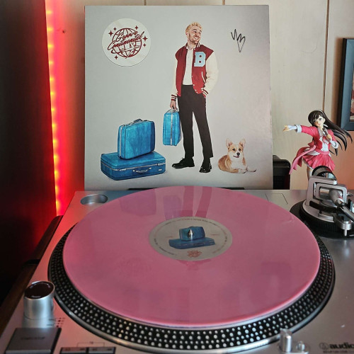 A pink vinyl record sits on a turntable. Behind the turntable, a vinyl album outer sleeve is displayed. The front cover shows Yung Bae standing with a piece of luggage in hand. There is more luggage stacked to the left of him, and a corgi laying on the floor to the right. It shows a logo that says Groove Continental in a Disco ball and plane. Yung Bae's autograph is in the upper right corner