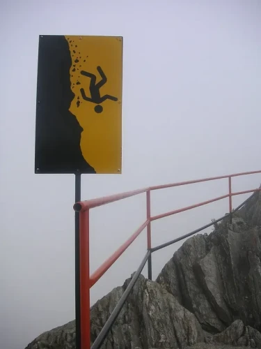 Warning sign posted atop a barrier protecting the edge of a steep cliff.
