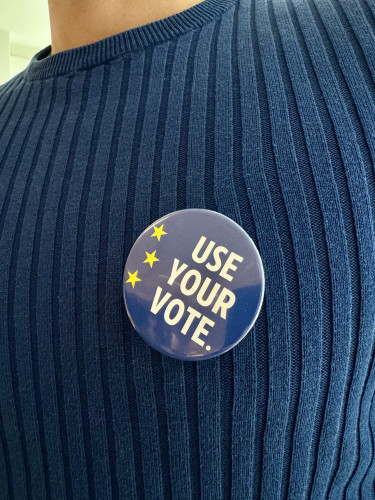 A person wearing a blue sweater. On it, a blue pin with part of the EU emblem and a white text which reads “Use your vote”. 