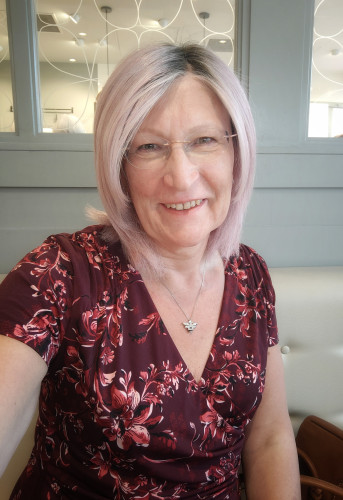 A pink haired woman sitting in a cafe wearing a burgundy, flower patterned v neck dress with a silver bee pendant necklace and silver hoop earrings.