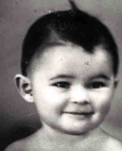 Black and white photograph of a girl's face. She is 2-3 years old. She has dark hair with a curl on top of her head. She has a round face and dark eyes. She is slightly smiling. Her right ear and exposed shoulders are visible.