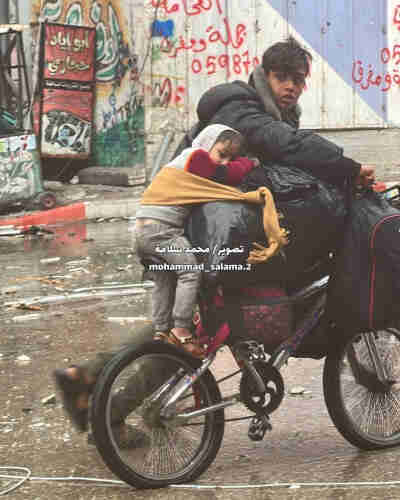 a palestinian teenager carrying all his belongings on his bicycle with child wrapped onto the luggage, probably moving to Rafah to get away from Israeli bombings