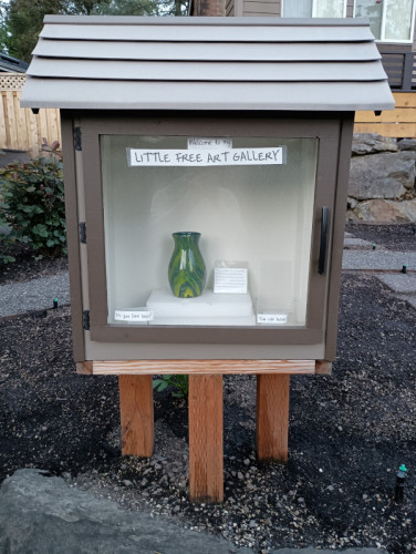 A brown wooden enclosure, about 3 feet tall by 2 feet wide and 1 foot deep, with a gray roof, and a big transparent door on the front. Inside sits a green and yellow swirled hand-painted vase which can be seen through the door. There is a sign on top of the door that reads "Welcome to my Little Free Art Gallery". There are two smaller signs on the bottom of the door which read "Do you like this?" and "You can have it".