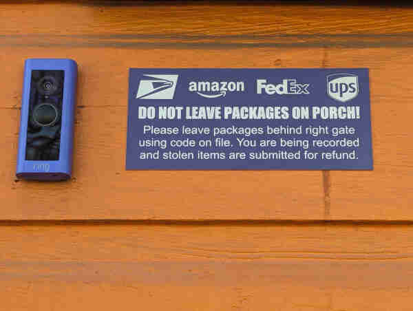A Ring camera mounted next to a sign on an orange-painted shiplap siding. The sign has the logos for USPS, Amazon, FedEx and UPS. It reads:

DO NOT LEAVE PACKAGES ON PORCH!!!
Please leave packages behind right gate
using code on file. You are being recorded
and stolen items are submitted for refund.
