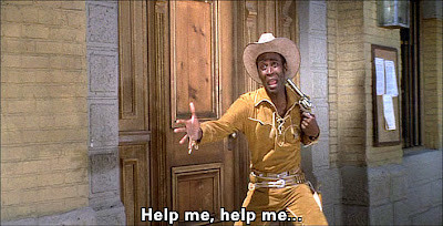 Still from "Blazing Saddles." The new sheriff is holding a gun to his own head, crying out, "Help me, help me!" 