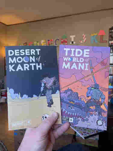 Two TTRPG books titled "Desert Moon of Karth" and "Tide World of Mani" are held up, featuring illustrated characters in otherworldly landscapes.