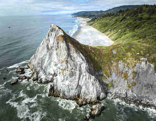 Color aerial photo taken from a drone of a white-cliffed headland that has a sharp peak extending out into the ocean with broken waves and sea foam at its base. Beyond is a light-colored beach with grassy sloped hillsides behind it. 