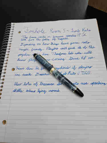 A picture of a notebook on a black table, with blue ink on it detailing the contents of a room in my upcoming supplement the Foxhole. A gray marbled fountain pen sits on top of it.