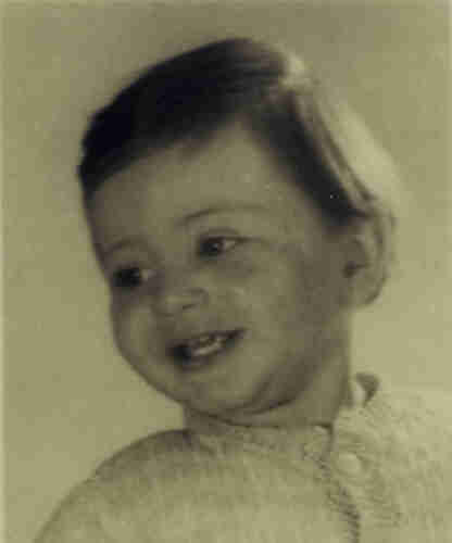 Portrait photograph of a young boy. He has blonde hair. His head is tilted to the left. He is smiling and his mouth is slightly open. He is wearing a white shirt buttoned up almost to the neck.