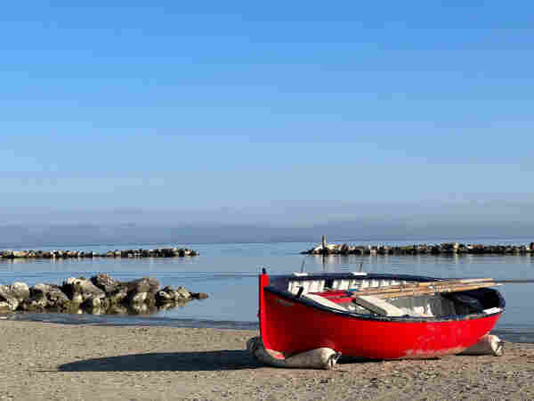 A vibrant red rowboat rests on a serene sandy beach, with its oars neatly placed inside. The calm waters of the sea gently lap at a rocky breakwater in the distance.  The clear blue sky overhead is devoid of clouds, reflecting the stillness of the scene, and hints of a distant fog blur the horizon line, creating a tranquil and picturesque setting.