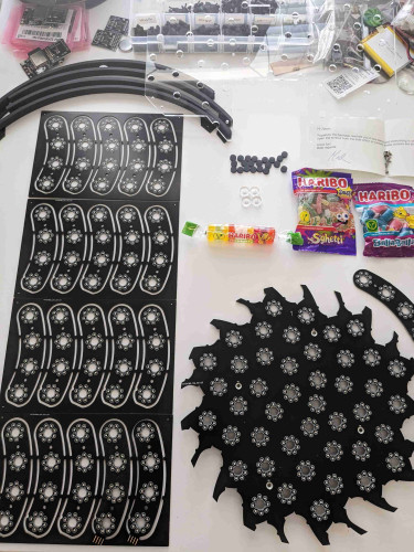 A photo of black PCBs laying on a white table. The PCB on the right is a jagged, sawblade looking disc with sharp points around the outside. It's covered in LEDs arranged into rings of eight, with the rings arranged into a Format spiral, like the center of a sunflower.
The PCBs on the left are arcs, with four of the same rings of eight LEDs on each arc. They're panelized, with five arcs per panel.
Also on the table are some Haribo brand gummy candies, some 3D printed parts, screws, etc.