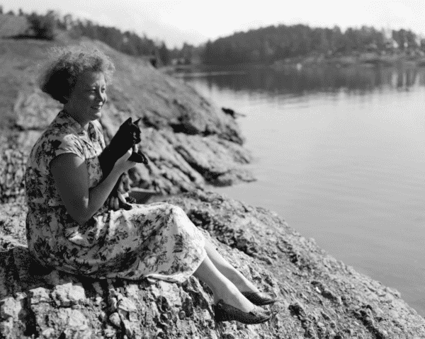Black and white photo of a white woman in knee length floor dress, sitting on a rock beach with a beautiful little shorthaired black kitten in her arms. The kitten looks brightly out over the water into the distance.