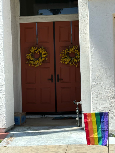 Smol pride flag in front of the entrance to a home, there are yellow wreathes on the doors and a white stucco exterior 