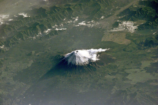 A photograph taken from the International Space Station shows the iconic snow-capped cone of Japan's famous Mt. Fuji in the middle of a seemingly flat, green forest. Several small puffs of cloud are in the sky, with one trailing off the mountain to the upper right. Ridges of smaller mountains are seen in the distance.