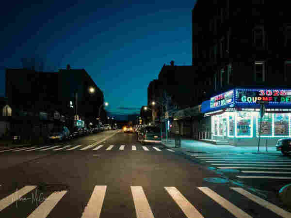 The view down a city street as the first hints of dawn peek above the horizon in the distance. Down the street, we see a car stopped in the middle of a lane with its brake lights glowing red. In the foreground on the right side of the street is a brightly lit corner store with neon lights proclaiming, "3026 Gourmet Deli". The rest of the street is still mostly dark, and this shop looks almost supernaturally bright by comparison.