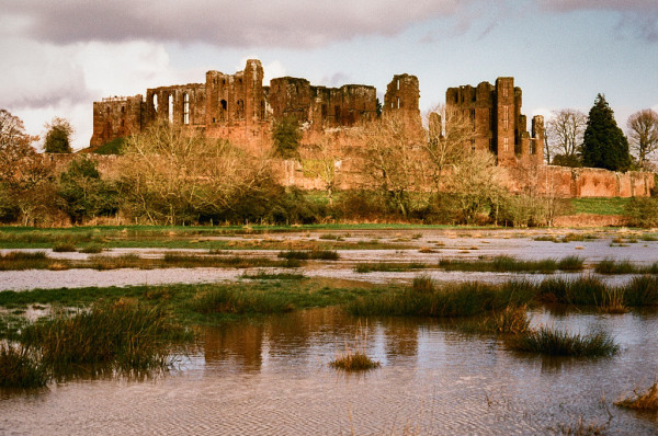A ruined stone castle spreads across most of the upper part of this photo, with dark clouds above it. A band of winter trees in front is lit by the afternoon sun. A flooded field lies in the lower half, with bunches of reeds poking through, and a reflection of the higher castle towers in the muddy brown water.