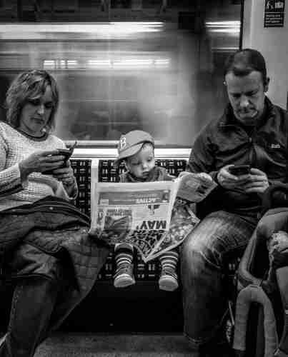 A shot of family on a train. While parents are on the phone, the kid is reading a newspaper 