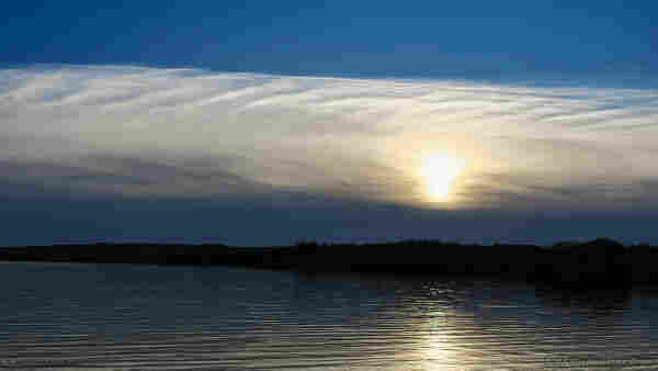 A photo of a body of still water with small ripples across the surface. A line of dark bushes and undergrowth is on the horizon. Above it is a bank of cloud with the sun visible behind and through it, and over that is a stretch of clear sky. The cloud is strangely plaited, with diagonal lines dropping from the top edge towards the left of the shot, and from the bottom edge down towards the right. It's oddly beautiful.