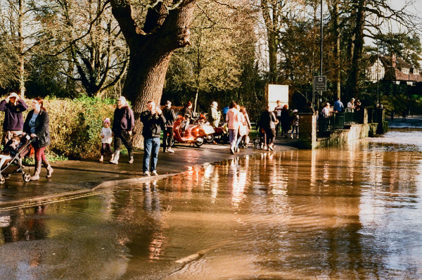 A highly saturated colour image of people gathering around the flooded ford, waiting for cars to splash through, or get stuck! Brown water in the bottom half of the frame, people milling about in the middle, and sunlit trees in the upper part.