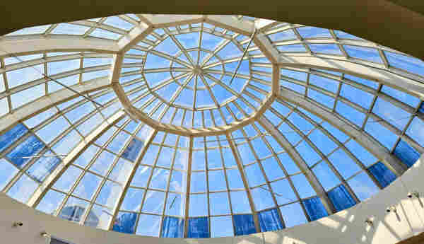 A large window Skylight in the ceiling of a central point in a shopping mall glows blue from sunlight and the clear blue skies outside.