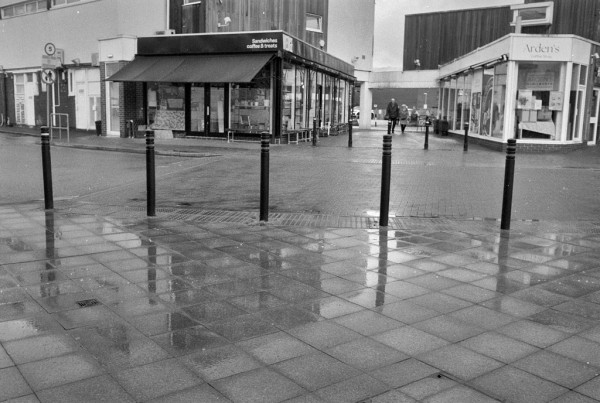 Damp paving slabs on the approach to a supermarket (guarded by metal posts) reflect the alley between two coffee shops, where  two people approach. Black and white photo.