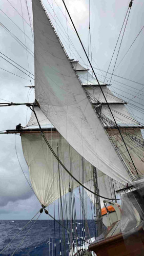 Person sitting on a heeling sail yard with all the square sails set and a stay sail in the foreground. 