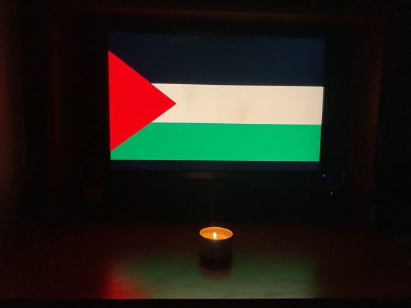 The Palestinian flag is displayed on a PC monitor in a darkened room. A candle is lit on the desk in front of it. 