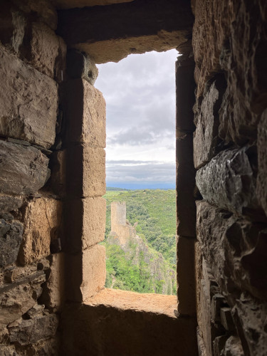 Photo of a narrow rectangular window opening in the stone wall of a castle tower, high on a limestone crag. Through the opening is a view over more castle ruins on a lower outcrop, a precipitous drop to the right. The steep hillsides are densely forested, continuing into the distance under a blanket of low cloud. Patches of washed out blue sky break up the cloud in the foreground.