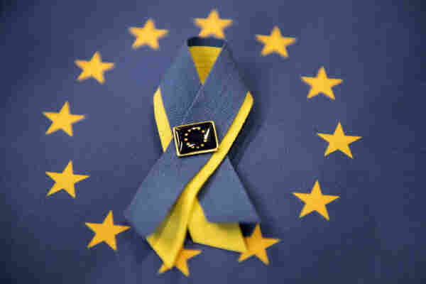 A photo of a ribbon with Ukrainian colours. The ribbon lies in the middle of the stars of an EU flag and has an EU pin on it.  