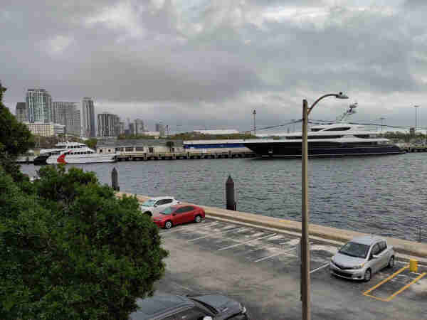 A large blue yacht, perhaps 175 feet (55 m) in length, to the right is docked in the Port of Saint Petersburg. To its left, a large ferry used to transport about 100 people across Tampa Bay is dwarfed by Penrod's yacht. In the background, the skyline of the city's condominiums is framed by a gray cloudy sky. In the foreground is the parking lot of my place of work with a few cars parked there.